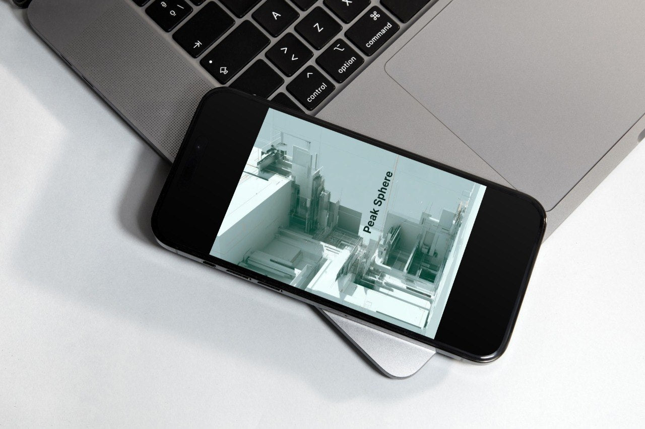A smartphone lying diagonally on a white surface, partially covering a silver laptop. The phone screen displays a presentation slide titled 'Peak Sphere'.